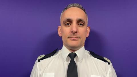 A man in a police uniform looks into a camera. A purple wall is behind him.