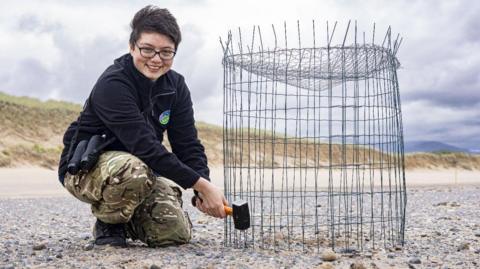 Steph Leow, wearing a fleece and camouflage trousers installing a bird cage on a stony beach.