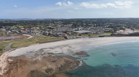 A sandy beach with blue coloured sea and rocks on left and housing behind with some areas of greenery.