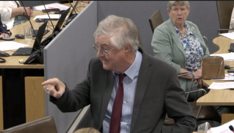 Mark Drakeford who has grey hair and glasses, wearing a grey suit jacket, shirt and red tie, pointing his finger and looking to the side with an angry expression. Behind him, sat nehind a wooden Senedd desk, is colleague Jan hut who also has short grey hair and glasses and is wearing a mint green blazer and blue and white floral blouse. She is looking at Drakeford with a concerned expression.