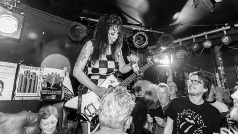 Woman in a monochrome checked shirt playing a guitar in front of a crowd of revellers