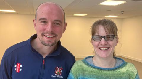 A man and a woman smile in front of a simple interior background. The man on the left is bald with stubble and is wearing a top that reads Colne Cricket Club. The woman has strawberry blonde hair, glasses, and a green and blue jumper