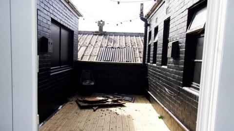 View form the smoking area it shows a low wall and wooden flooring with with black painted walls on either side in the background is the roof of another building