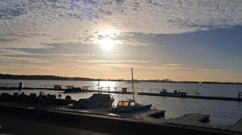 Two boats are moored up at a pontoon in the foreground of the picture. Behind them an expanse of water stretches to the horizon. The sky is still blue but darkening with a hazy setting sun in its centre.