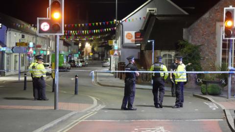 A road at night in Hailsham town centre with police officers standing with back to camera and police tape across road