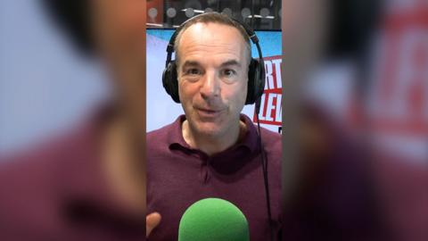 Martin Lewis is sat in front of a green mic with black headphones on. He is mid speech.
