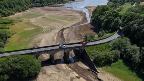 A drone view shows vehicles passing over a bridge across a dry section of the Lindley Wood Reservoir, which has been largely drained.