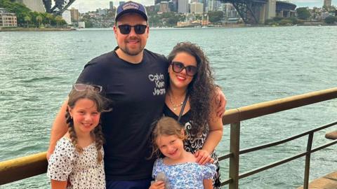 Harry, Brydee, Eliza and Lillie are standing outside on holiday. They are near water, with a railing behind them, and they are wearing summer clothes and looking happy.