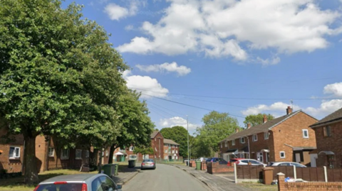 There are houses on either side of the street, some with cars parked outside. There are a couple of large trees on the left side of the road.