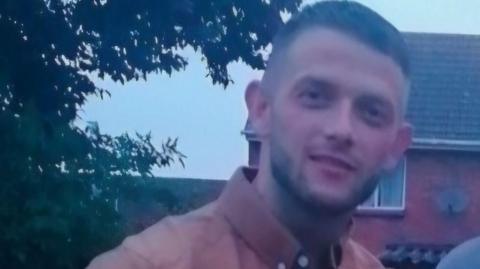 A slightly blurred photo of Danny Fowler, who is smiling for the camera and wearing a coral shirt. A house can be seen in the background.