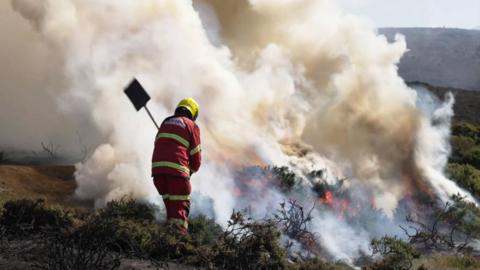 A fireman attends a wildfire in North Wales. Smoke can be seen billowing from a burning moor and a fireman with a shovel is in the foreround