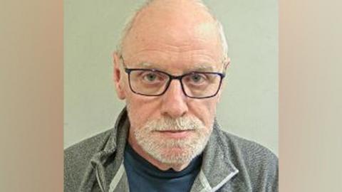 Macdonald's mugshot. He was very short white hair, balding on top nd a short white heard. He has black-framed glasses and is wearing a navy tshirt under a grey zipped top