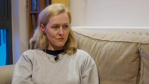 A woman wearing a light grey sweatshirt sits on a sofa in a living room, looking to one side during an interview.