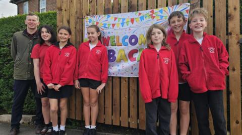 Six students and the head teacher are standing in front of a sign that reads 'welcome back. 