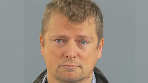A police custody photo of Leo Norman against a grey background. He has light brown hair which is curly on top, blue eyes and a short goatee beard. He wears a jacket with the collar turned up.