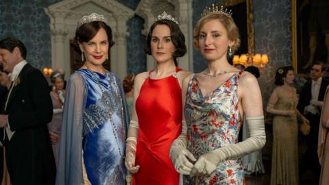 Three female characters from Downton Abbey stand together looking at the camera. From left to right Cora, Lady Mary and Lady Edith. Cora wears a diamond tiara with a blue velvet ball gown, Lady Mary wears a red strappy silk gown with a diamond tiara and Lady Edith wears a silk strappy floral gown with a diamond tiara.