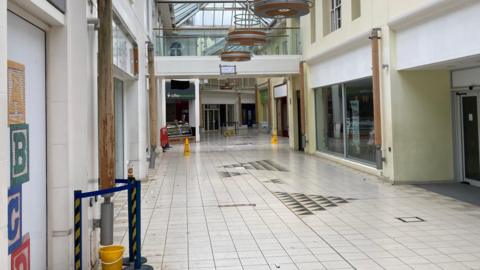 The inside of shopping centre - showing white flooring and several retail units which are empty.
