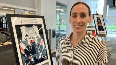 Weightman stands beside a framed display that showcases a white sports jersey with the text "GREAT BRITAIN". The jersey includes the Adidas logo and the British Olympic Association logo, suggesting its connection to Team GB. The display also contains a photograph of an athlete in action and a plaque. In the background, another similar framed display is visible on an easel.