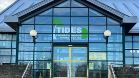 a glass-fronted building with a pitched roof, with Tides Leisure and Indoor Tennis Centre written above the door