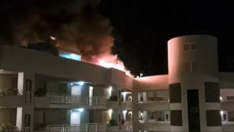 Flames and smoke pour from the top of a multi-storey white building at night.