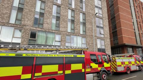 A fire and rescue service truck parked outside a tall accommodation block that is grey and brown with pale green curtains. There is a smaller truck in front. 