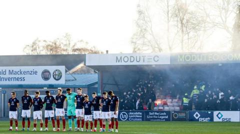 Ross County players stand in a line for the minute's silence. Behind them smoke from flares rises from the Jail End stand. Football fans and stewards look on.