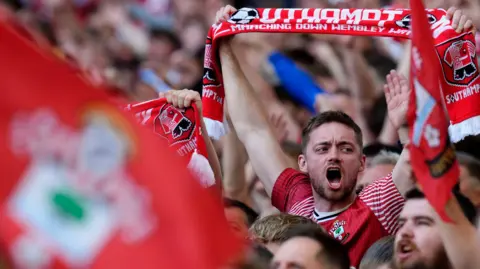 Southampton fans cheer their team on in the stands at Wembley in 2026. In the foreground is an out-of-focus flag, red in colour, with a Southampton FC badge on. In focus fans in Saints shirts cheer and hold aloft red scarves.