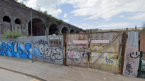 Metal fences covered in graffiti and a metal gate with no parking notice on it with a viaduct in the background