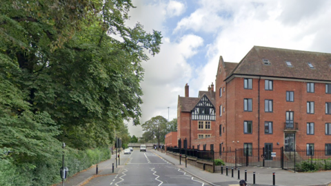 A single carriageway road with green trees to the left and tall brick buildings on the right