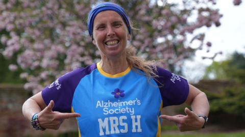 Apryl wearing a light blue running vest with Alzheimer's Society written on the front - the charity she's running in aid of at Sunday's London Marathon. Her top also says Mrs H. She's smiling and pointing to her name on her vest.