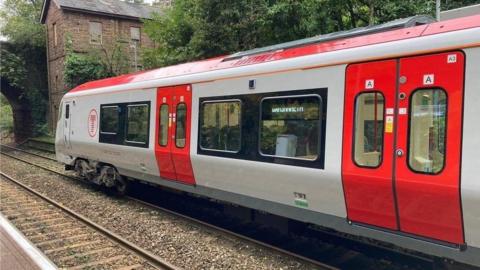 A grey and red train in Transport for Wales colours, running on dual railway tracks near a bridge pictured in the distance on the left hand side.