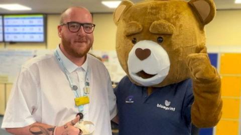 A man wearing black glasses and a white shirt with a light blue lanyard and badges stands next to a bear mascot. The mascot wears a dark blue T-shirt that says "blueprint" on it, in front of a yellow wall. Screens can be seen in the background.