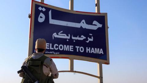 A rebel fighter stands next to a sign saying "Welcome to Hama" on the Damascus-Aleppo highway, Syria (3 December 2024)