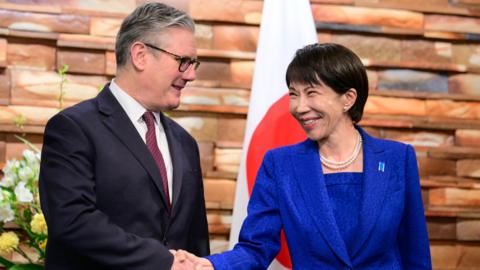 Keir Starmer and Sanae Takaichi wearing suits, shaking hands, and smiling in front of a Japanese flag and flowers