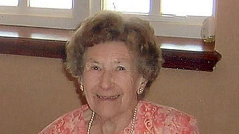 Una Crown: She is smiling at the camera while sitting in front of a beige-coloured wall and window. She is wearing a pink patterned dress with a pearl necklace and shiny earrings.