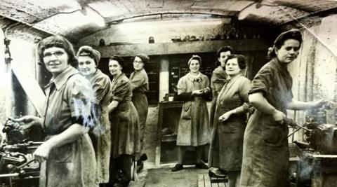 A group of women work in a makeshift munitions factory beneath the arcade in New Brighton, Merseyside. They are dressed in work overcoats and have their hair tied up or under headscarves as they operate machinery making bullets and shell casings. 