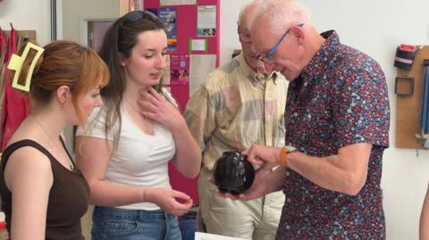 A group of 4 people two men and two women looking over a tea pot whilst discussing how it was made.