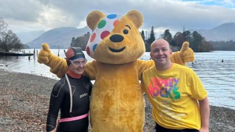 Pudsey Bear, who is yellow and has a spotty-coloured white bandage over one eye, standing in the middle of Joanne McKenzie, who is wearing a black wetsuit, and James Philips in a yellow 'Pudsey Rocks' T-shirt. Derwentwater can be seen behind them.