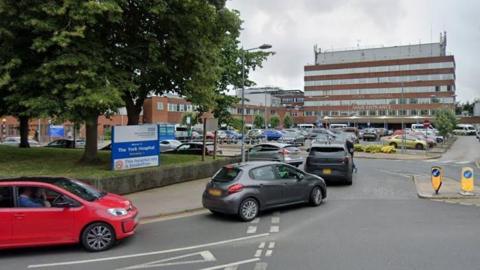 The entrance area to a large hospital complex on an overcast day. In the background, there is a multi-storey hospital building with horizontal red and white stripes across its façade. On the front of this building, the words “YORK HOSPITAL” and “MAIN ENTRANCE” are clearly visible in white lettering.
In the foreground, a busy road leads into a car park area. Several vehicles, including cars and vans, are either driving toward the entrance or parked in the lot. A small traffic island with two yellow-and-blue directional bollards sits in the middle of the road, guiding vehicles as they turn.