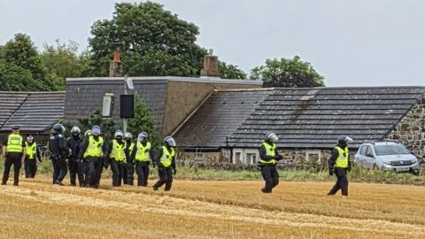 Ten police officers wearing helmets standing in a field outside a row of properties. 