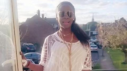 Maureen Rickards standing next to a French window. She is wearing a white top with sheer sleeves, aviator sunglasses and a lot of jewellery.