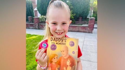 Isabella is holding her book and smiling at the camera. The front cover reads: Daddy's little monsters. There is a drawing of Isabella cuddling her dad Chris and her younger sister. There are three purple fluffy balls to represent the illness. 