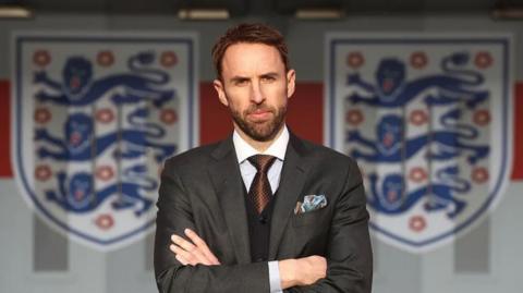Gareth Southgate standing with his arms crossed in front of England’s Three Lions Crest