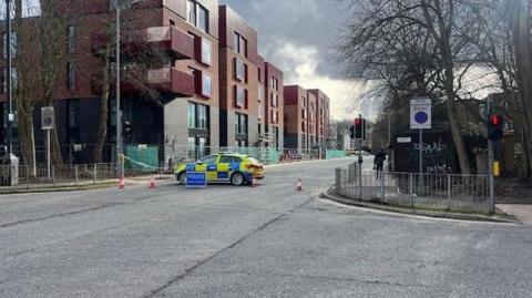 A shot of a street crossing, there is a police care across the road, as well as cones and police tape blocking the left exit off the road. To the far left there are buildings, with red brick at the top and black bricks at the bottom.