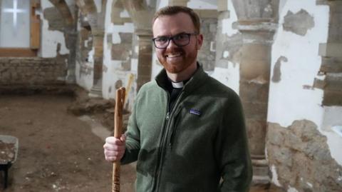 The Revd James Shelton, pictured from the waist up, with short reddish hair and a close-cropped beard, wearing spectacles, a dog collar and a dark green zipped up fleece, and holding a staff in his right hand. He s standing in a crypt with arched vaults of exposed stone, and the walls between the pillars are semi-plastered. At the far end is a window with a cross etched in the opaque glass. Building work is taking place as evidenced by the exposed floor, a wheelbarrow and a hose.