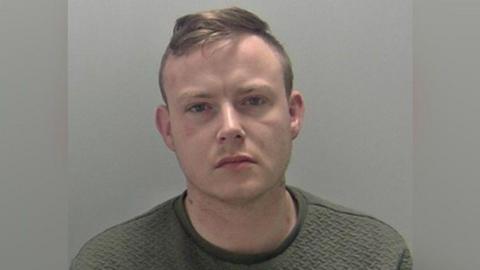 A mugshot of a man with short brown hair that is in a quiff. He is wearing a dark grey jumper and staring into the camera with a straight face. 