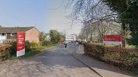 The entrance to a school site. There are red signs either side of a driveway, and barriers on entrance to a car park.