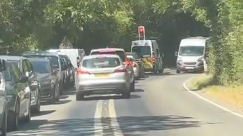 A line of cars parked on a road with white lines down the middle. There are trees and hedgerows at the side of the road. There is a set of traffic lights in the distance and vehicles travelling in both directions.