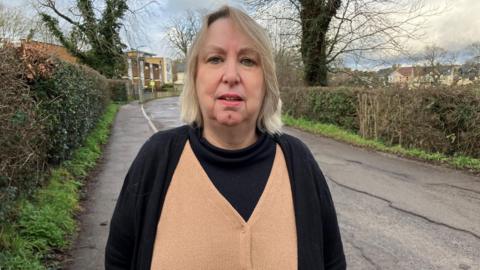 Nicky Brownjohn stood on Princes Road, Earlswood, Surrey. She has blonde hair and is wearing a black cardigan and tan top. 