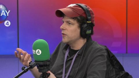 Jesse Eisenberg wearing red cap and headphones speaking into a mic in radio studio